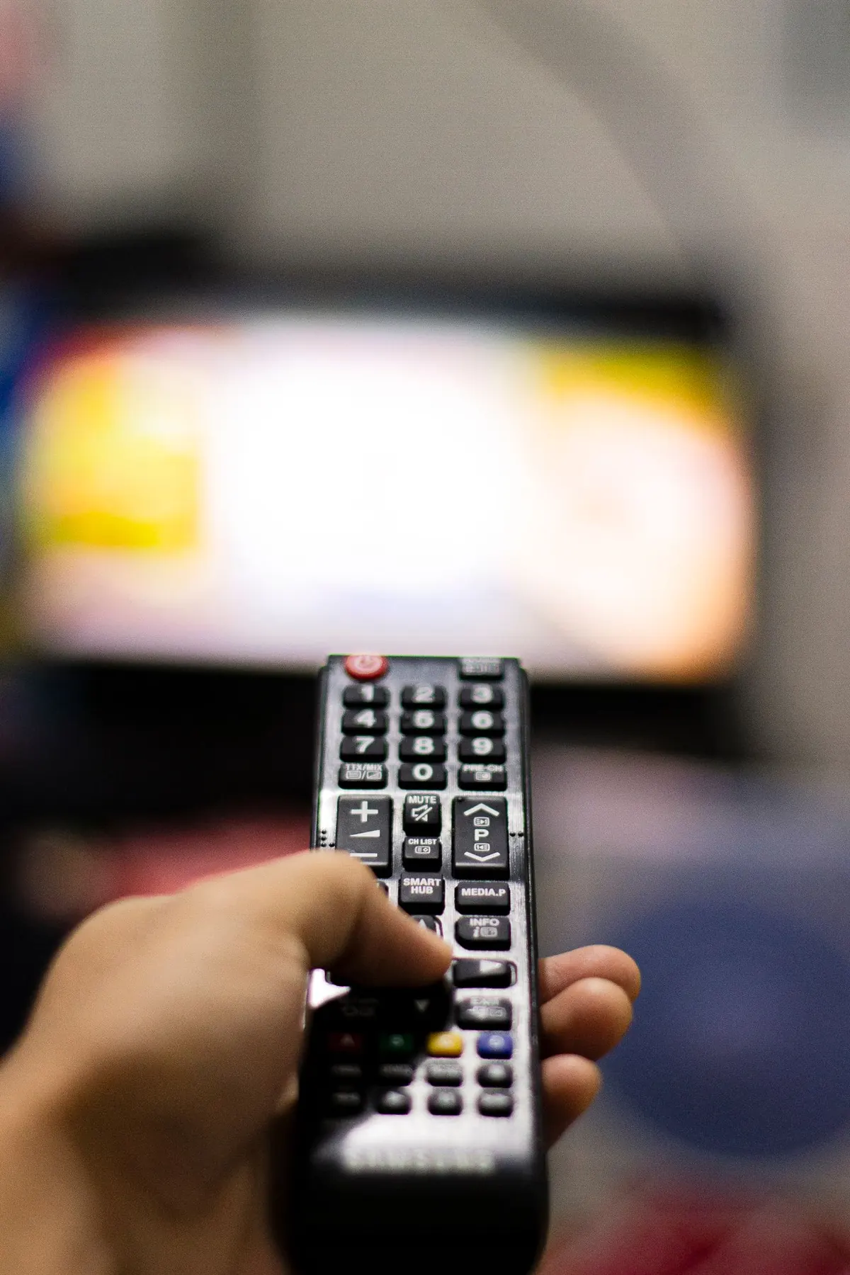 A hand holding a TV remote with a television in the background